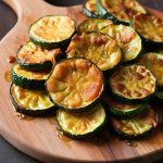 Golden brown zucchini slices pan-fried to crispy perfection, arranged on a white plate and garnished with fresh herbs. The zucchini slices have a crunchy exterior and tender interior, served as a delicious and simple appetizer or side dish.